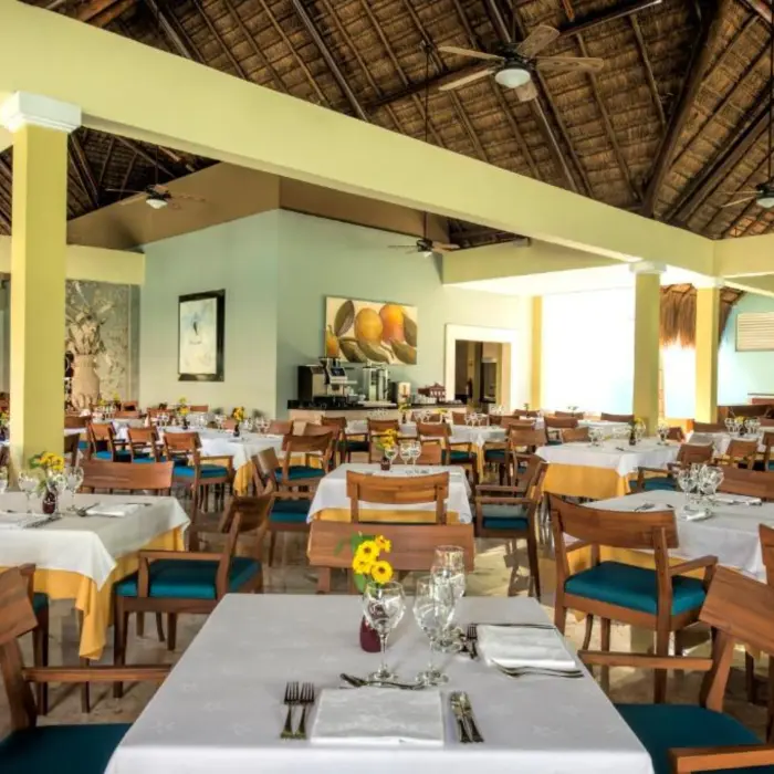 An empty restaurant with many tables and chairs set up, a thatched roof, and large windows.