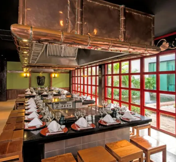A modern restaurant with a long table, chairs, and a large hood over a cooking station.