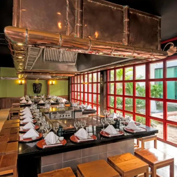 A modern restaurant with a long table, chairs, and a large hood over a cooking station.