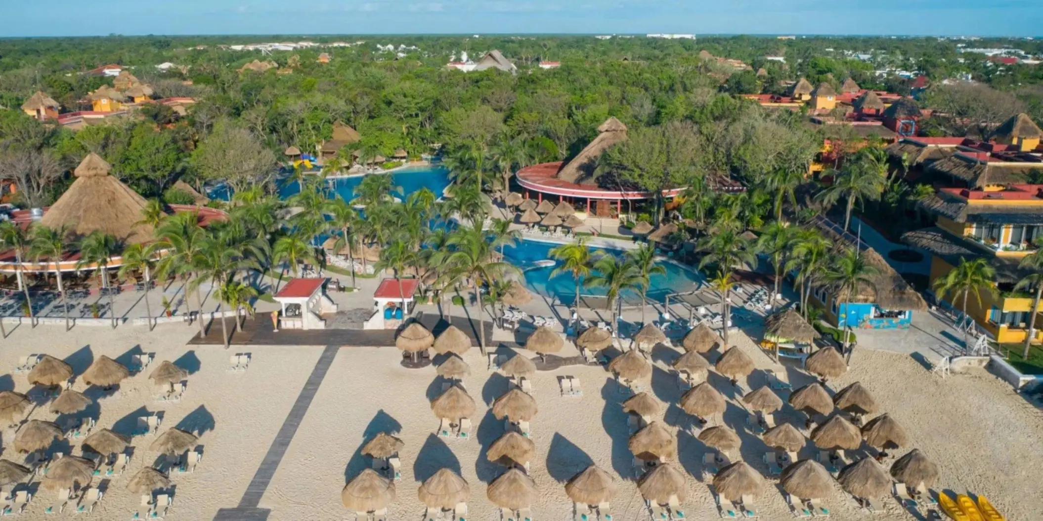 An aerial view of a resort with multiple pools, thatched-roof buildings, and a wide sandy beach with umbrellas.