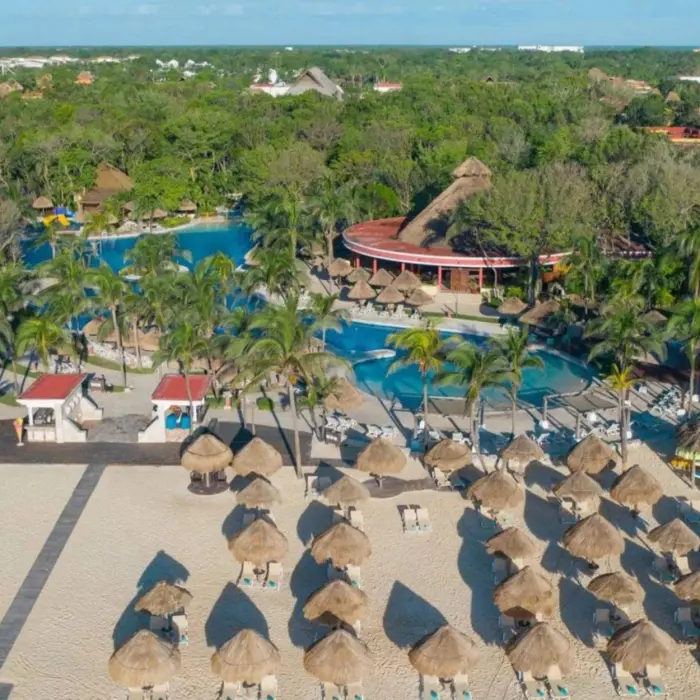An aerial view of a resort with multiple pools, thatched-roof buildings, and a wide sandy beach with umbrellas.