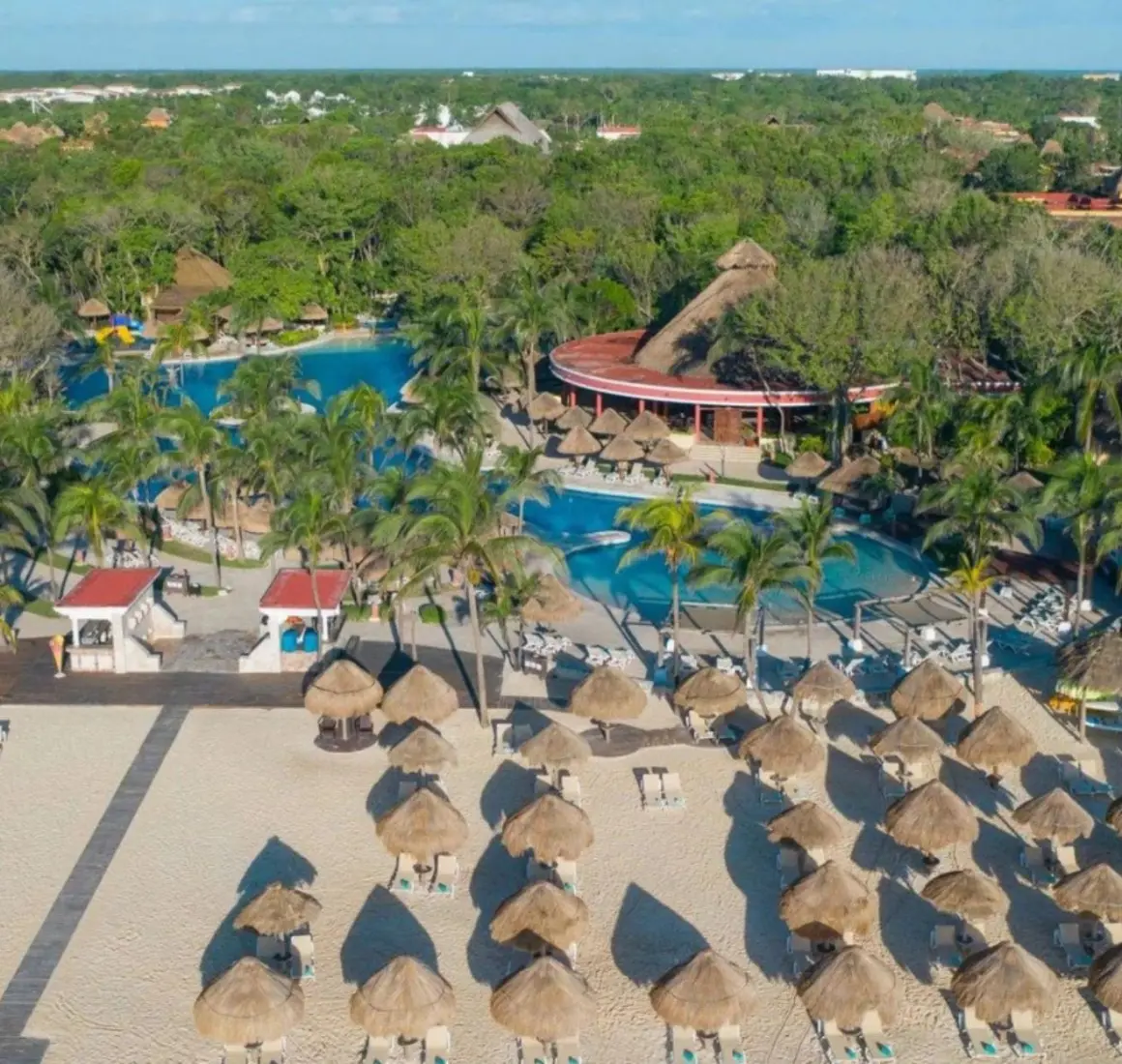An aerial view of a resort with multiple pools, thatched-roof buildings, and a wide sandy beach with umbrellas.