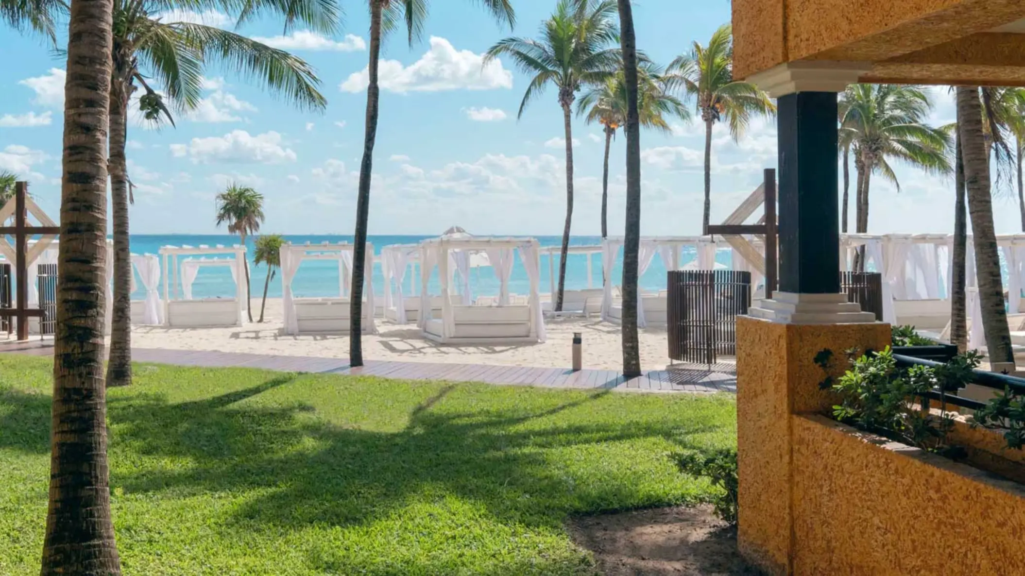 White cabanas on a sandy beach, with lush green grass and palm trees leading to the blue ocean.