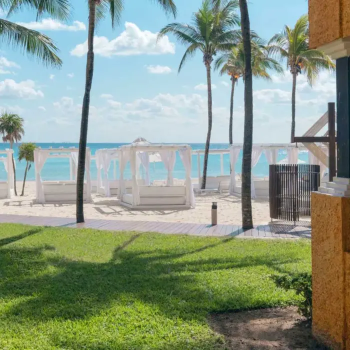 White cabanas on a sandy beach, with lush green grass and palm trees leading to the blue ocean.