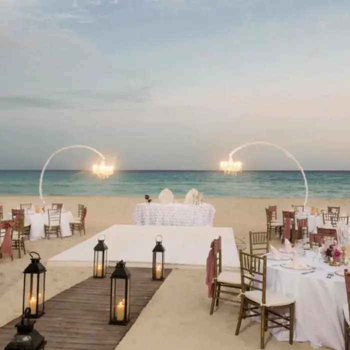 An elegant beach wedding or event setup with white tables, chairs, and chandeliers by the ocean.