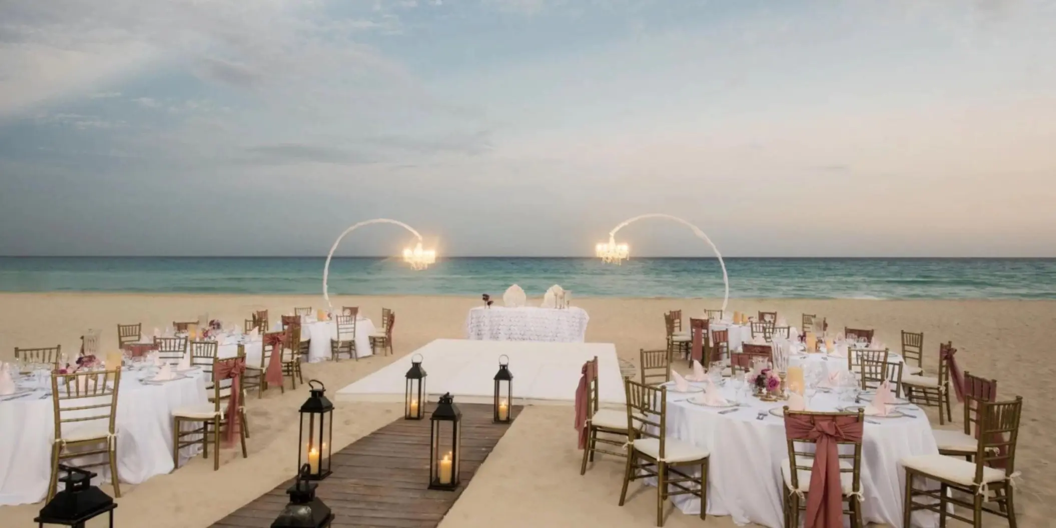 An elegant beach wedding or event setup with white tables, chairs, and chandeliers by the ocean.