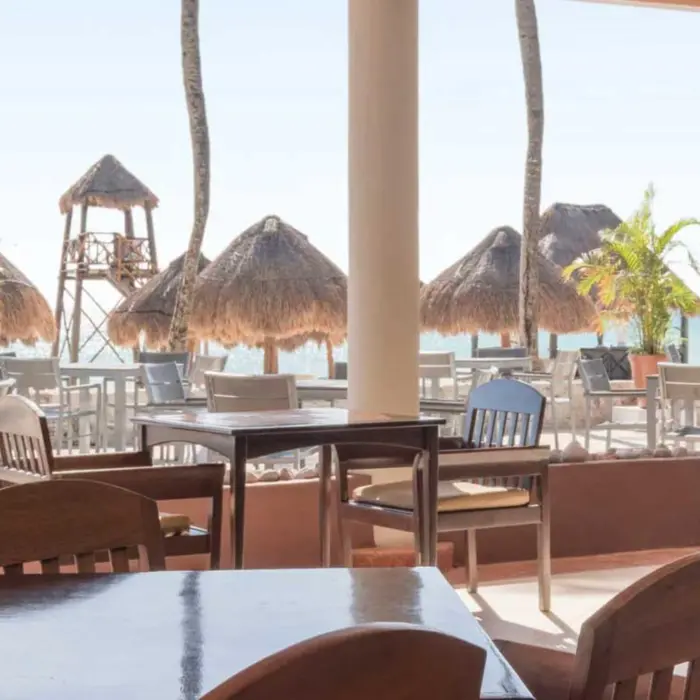 An outdoor restaurant with tables and chairs, offering views of the sandy beach and ocean with palm trees.