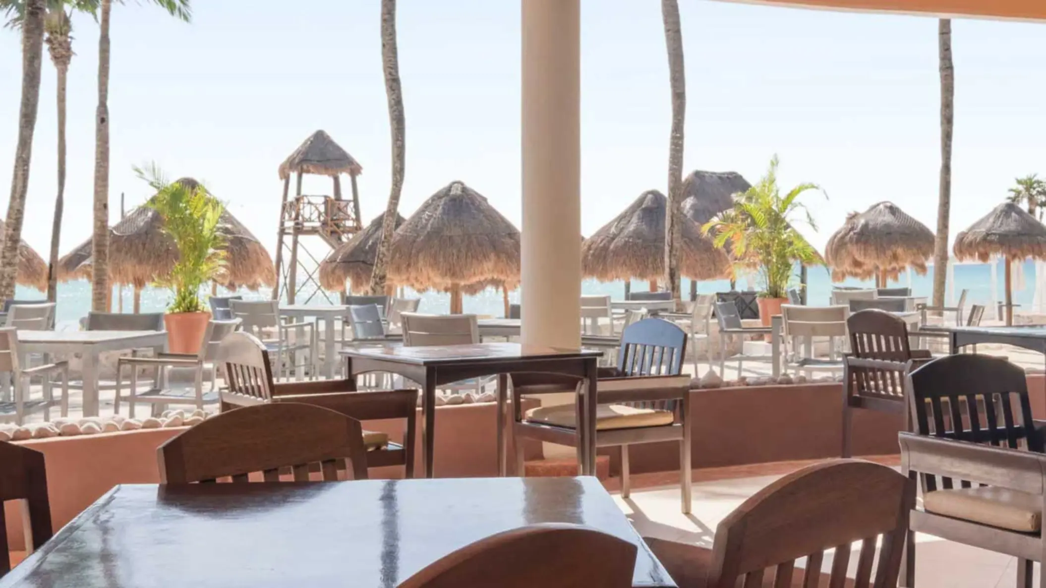 An outdoor restaurant with tables and chairs, offering views of the sandy beach and ocean with palm trees.