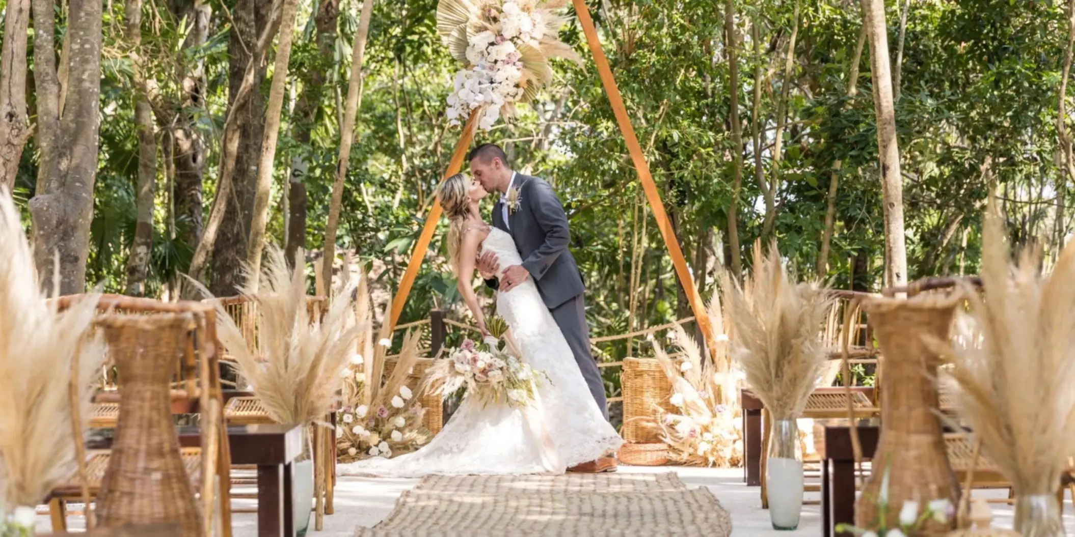 A bride and groom kissing under a decorated arch in a lush jungle setting, surrounded by natural elements.