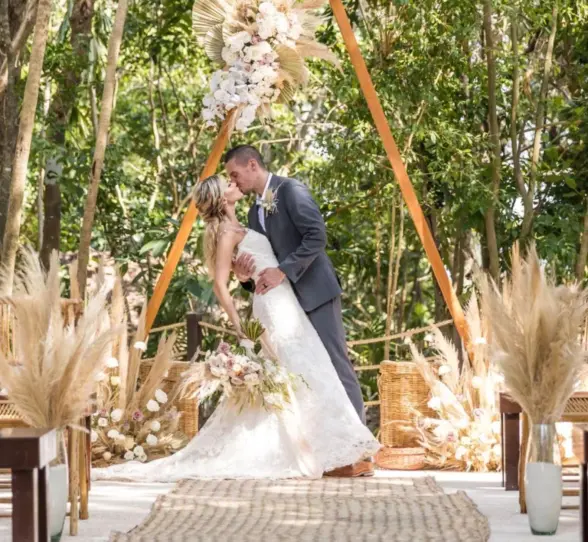 A bride and groom kissing under a decorated arch in a lush jungle setting, surrounded by natural elements.