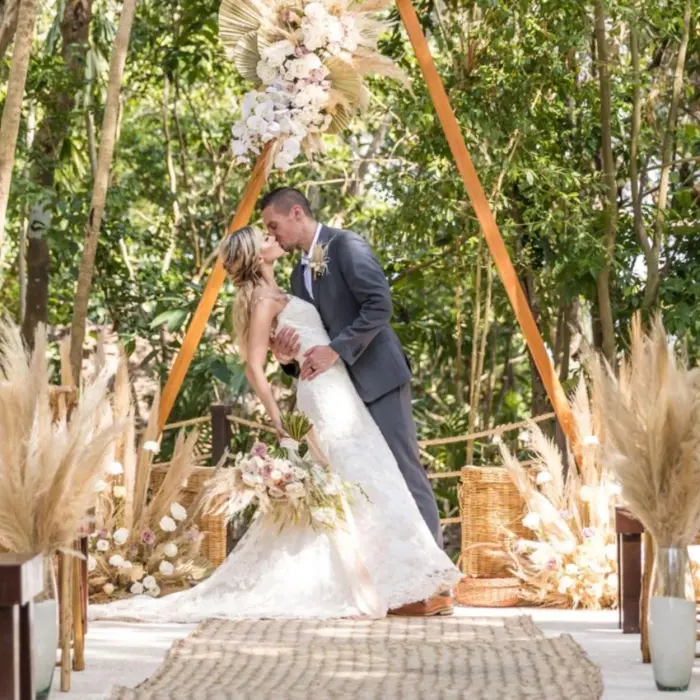 A bride and groom kissing under a decorated arch in a lush jungle setting, surrounded by natural elements.