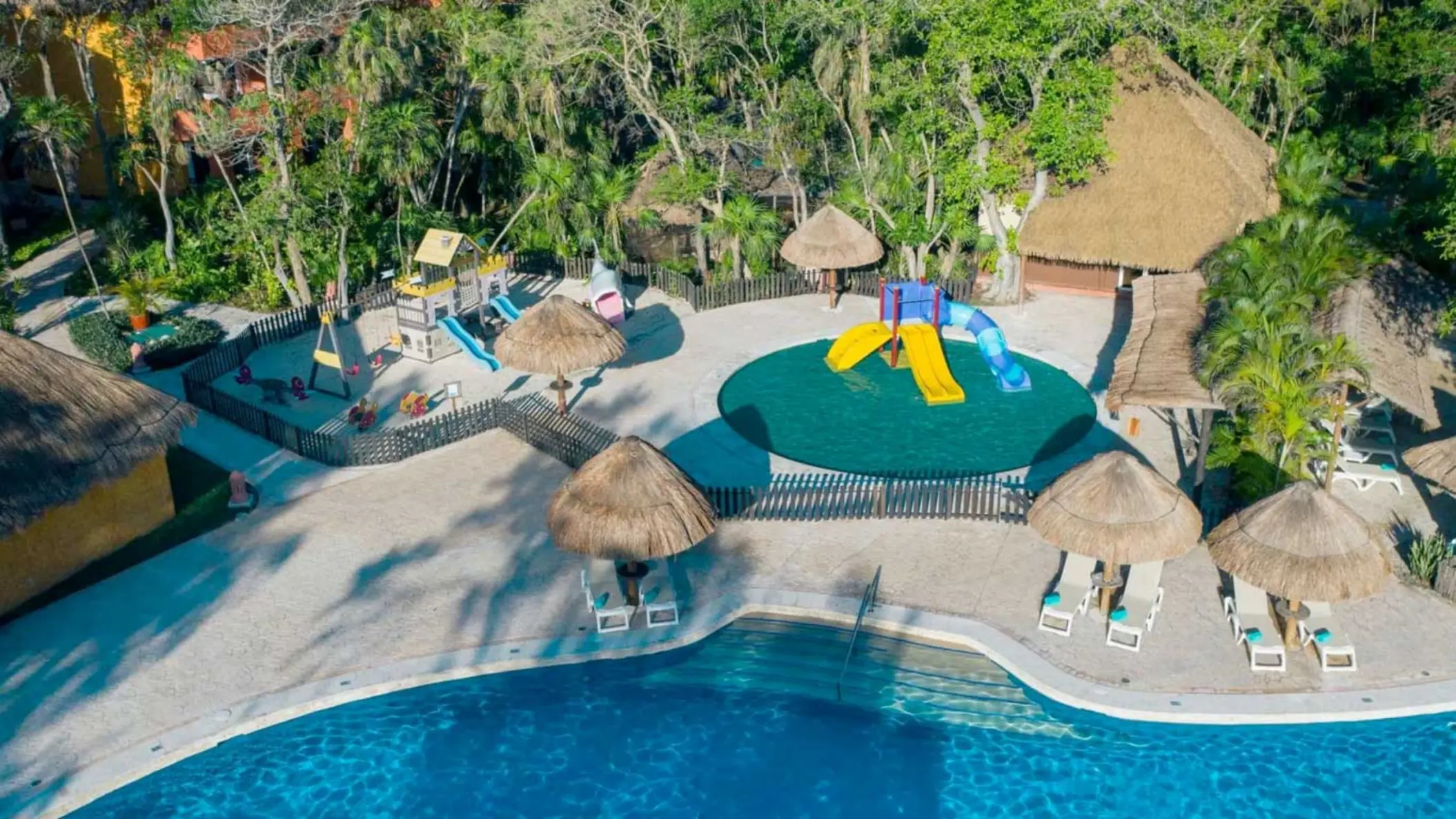 An aerial view of a resort pool next to a colorful children's playground with slides and swings.