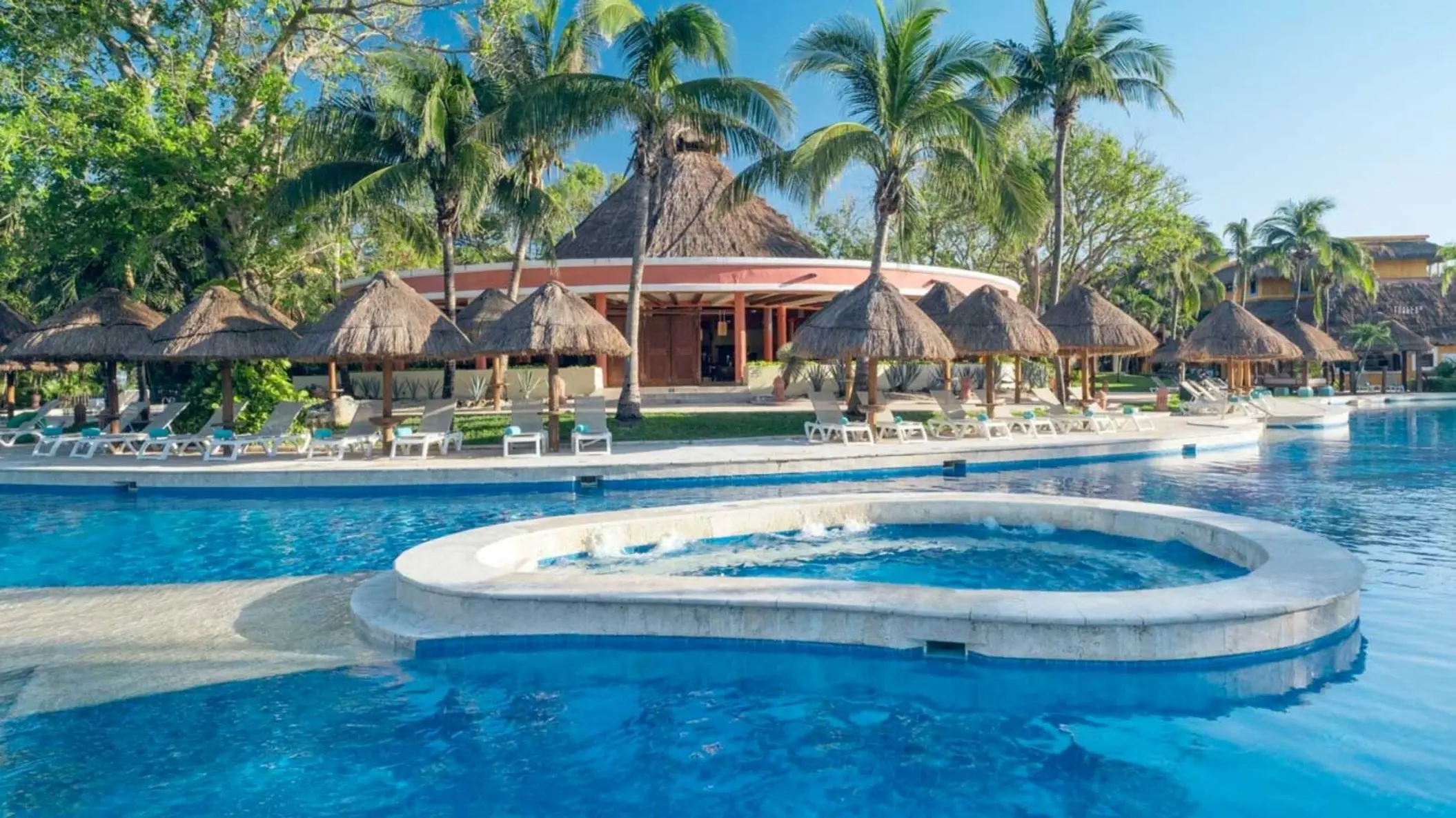 A large, inviting swimming pool with palm trees, thatched-roof structures, and lounge chairs.