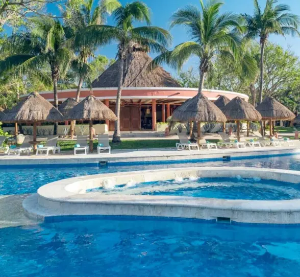 A large, inviting swimming pool with palm trees, thatched-roof structures, and lounge chairs.