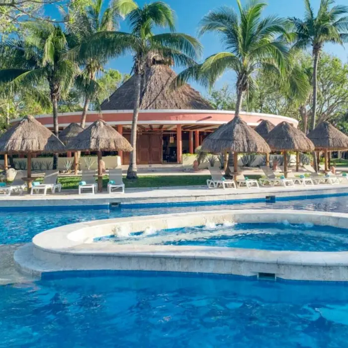 A large, inviting swimming pool with palm trees, thatched-roof structures, and lounge chairs.