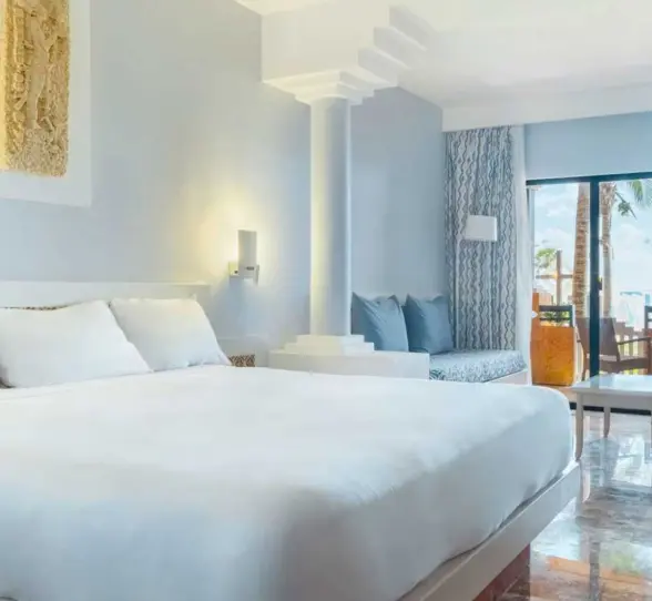 A light-colored hotel suite with a king-size bed, a sitting area, and a balcony overlooking the ocean.