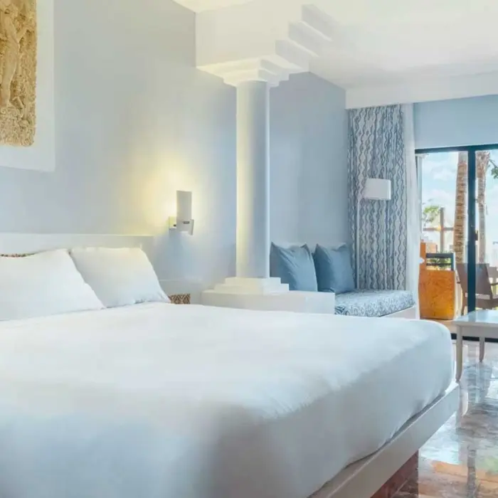 A light-colored hotel suite with a king-size bed, a sitting area, and a balcony overlooking the ocean.