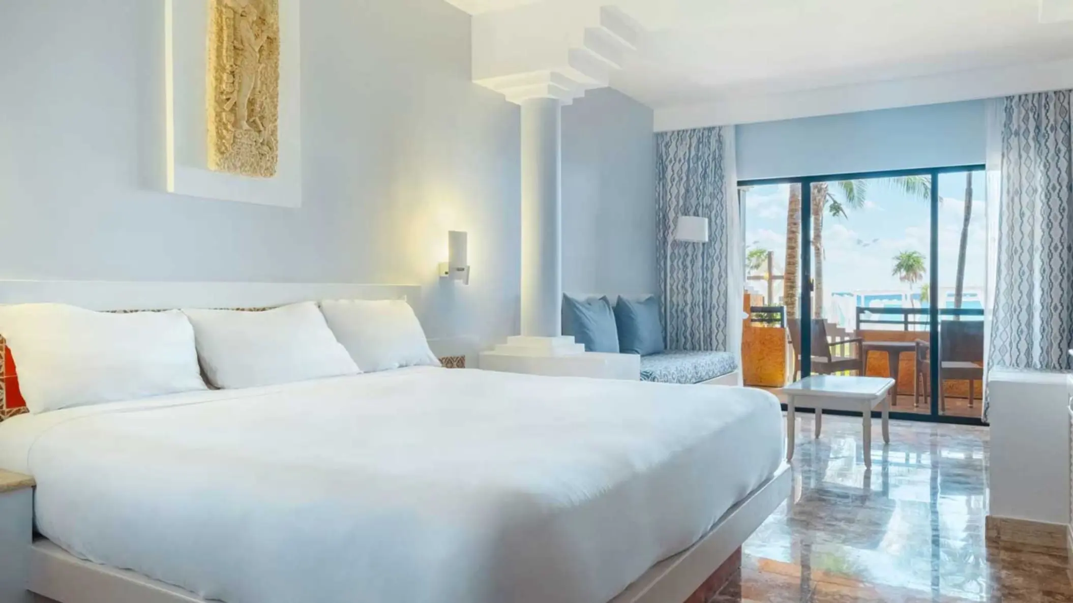 A light-colored hotel suite with a king-size bed, a sitting area, and a balcony overlooking the ocean.