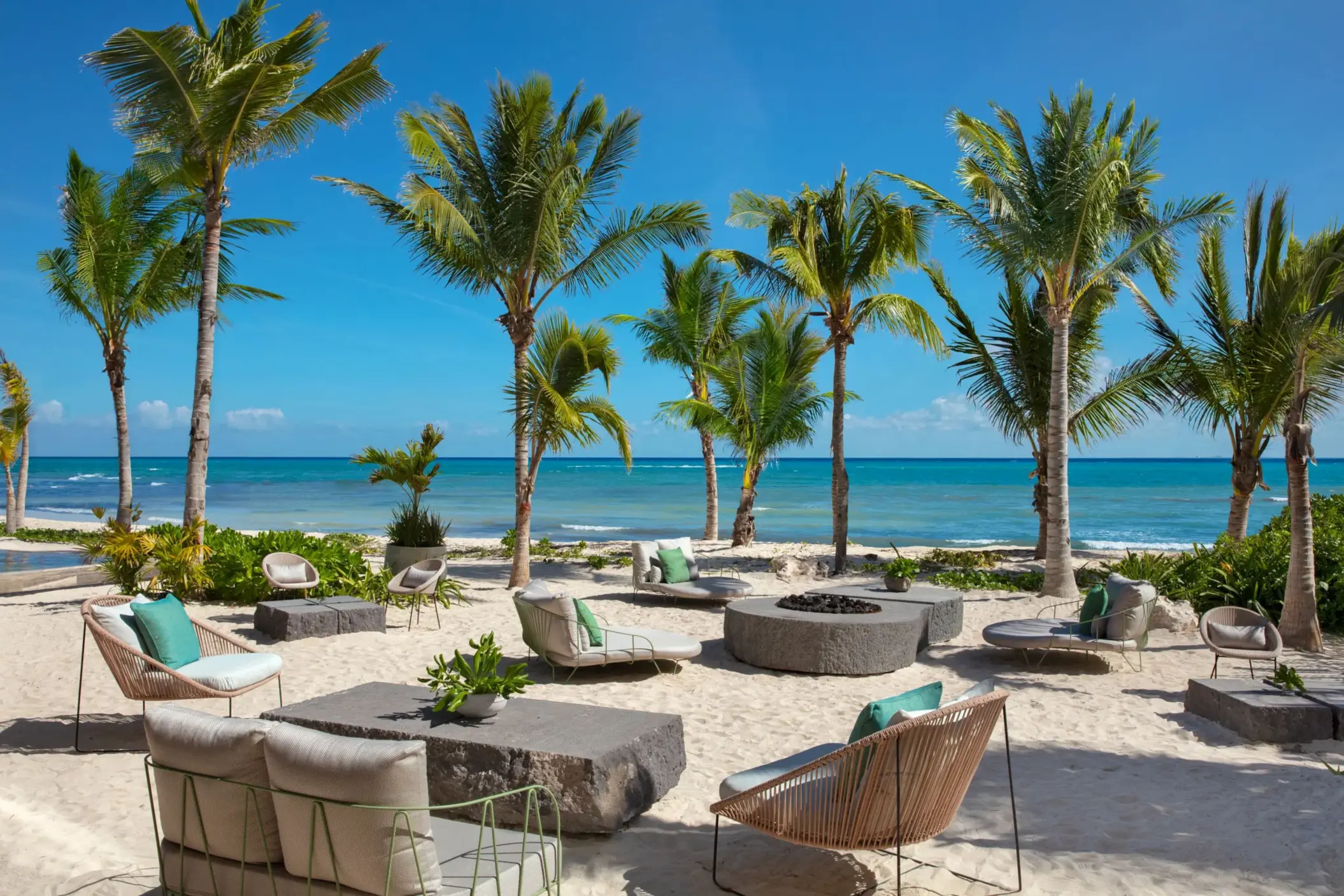 Chic beach lounge area with modern chairs and firepit surrounded by palm trees.