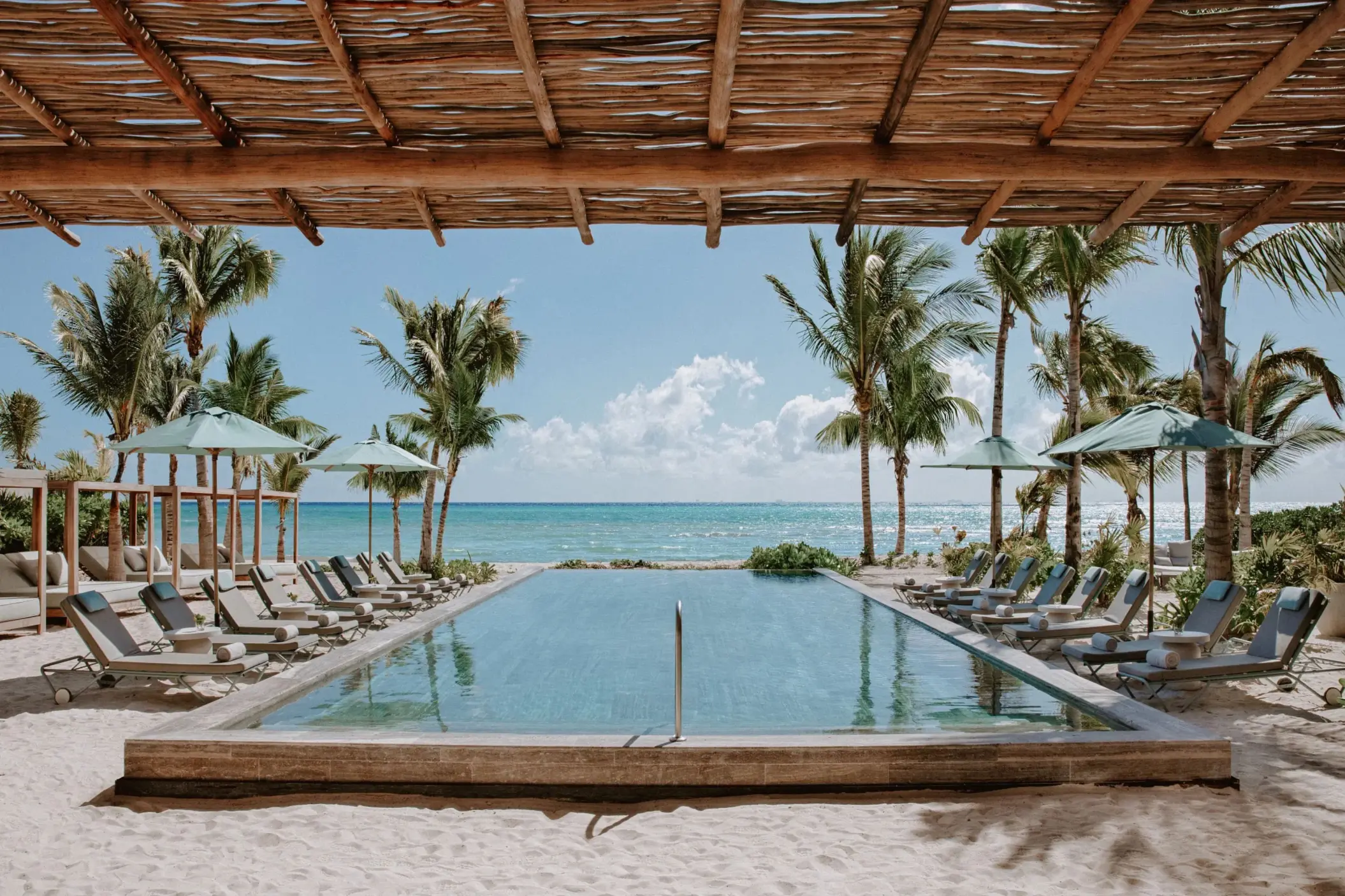 Beachfront pool surrounded by loungers and palm trees under a wooden pergola.