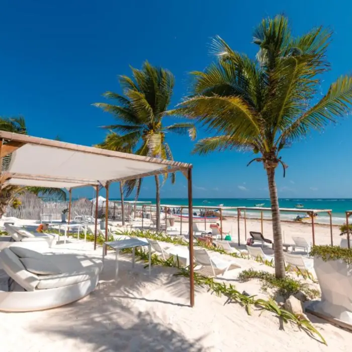The vibrant beach club at Kai Tulum Hotel with white cabanas, lounge chairs, palm trees, and the ocean.