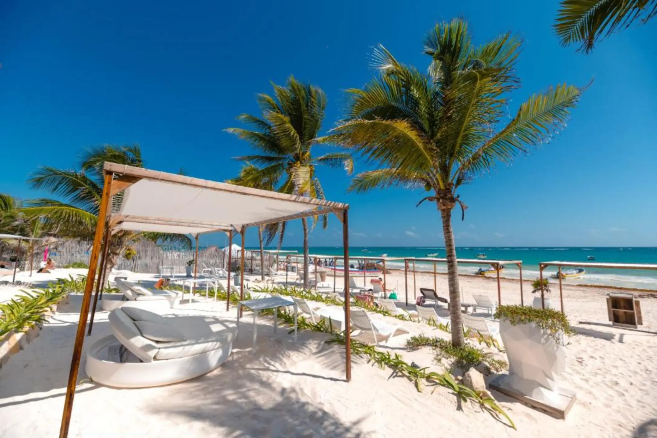 The vibrant beach club at Kai Tulum Hotel with white cabanas, lounge chairs, palm trees, and the ocean.