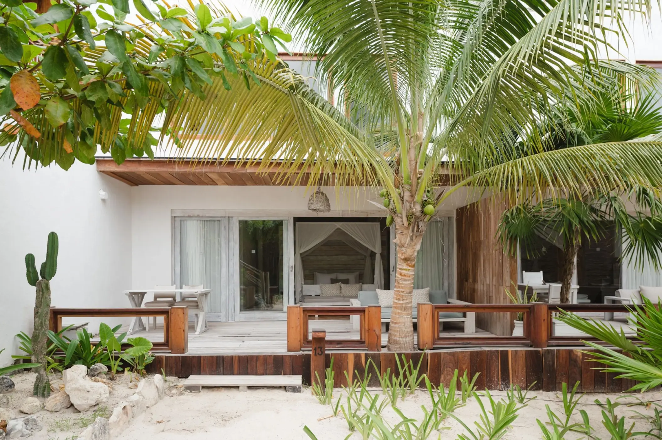 The exterior of a Garden View Suite at Kai Tulum Hotel, featuring a wooden deck, tropical plants, and sand.
