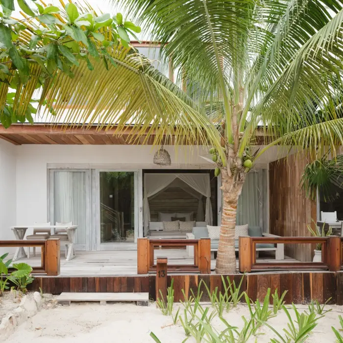 The exterior of a Garden View Suite at Kai Tulum Hotel, featuring a wooden deck, tropical plants, and sand.