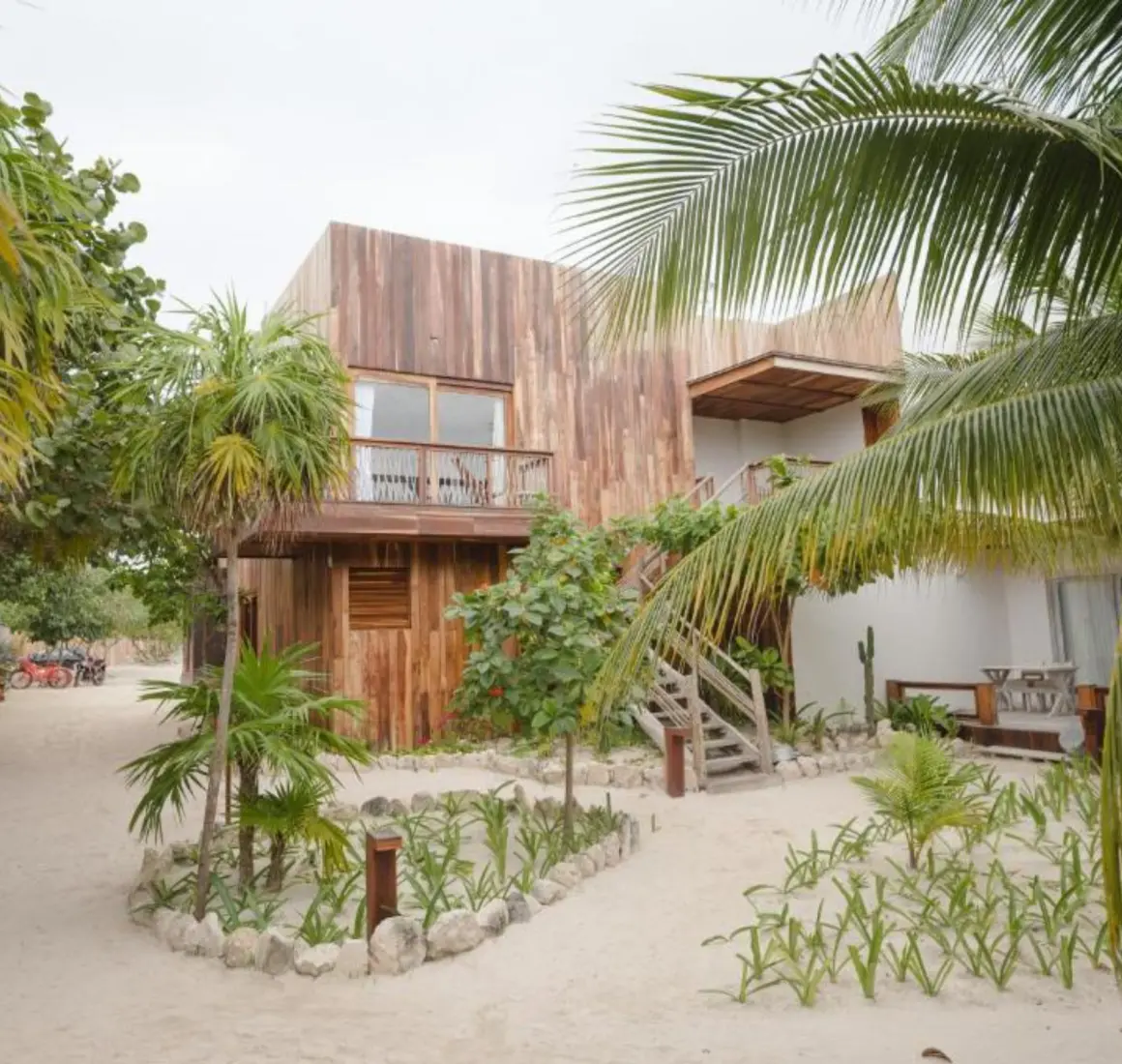 Exteriors of the suites at Kai Tulum Hotel, with wooden facades, balconies, and sandy pathways amidst lush plants.