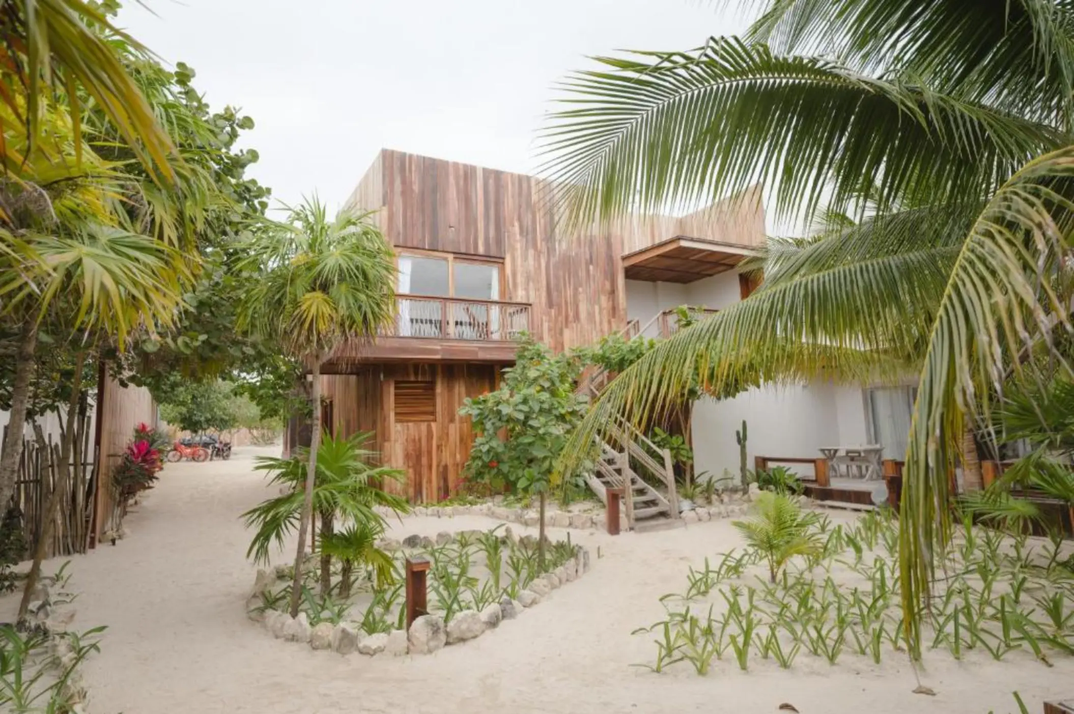 Exteriors of the suites at Kai Tulum Hotel, with wooden facades, balconies, and sandy pathways amidst lush plants.