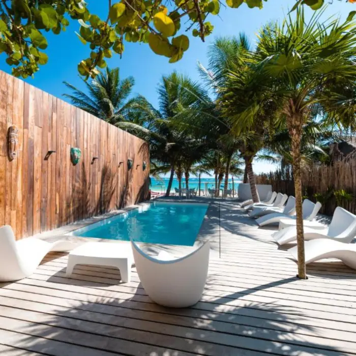 A serene swimming pool at Kai Tulum Hotel, surrounded by white lounge chairs, palm trees, and a wooden deck.