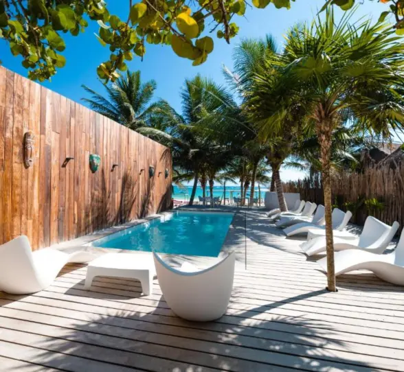 A serene swimming pool at Kai Tulum Hotel, surrounded by white lounge chairs, palm trees, and a wooden deck.