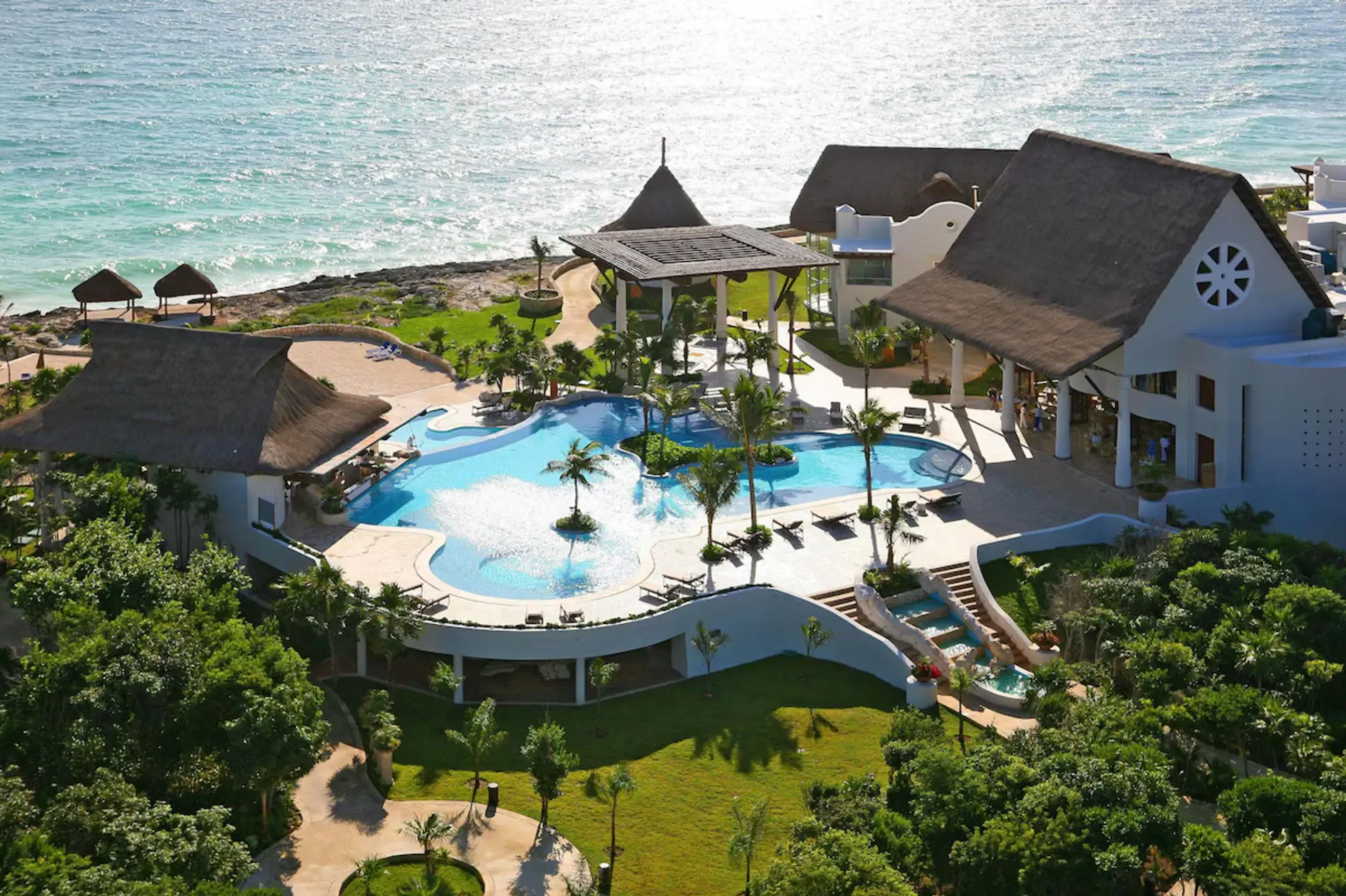 An aerial view of a resort with a large pool, lush landscaping, and buildings next to the ocean.