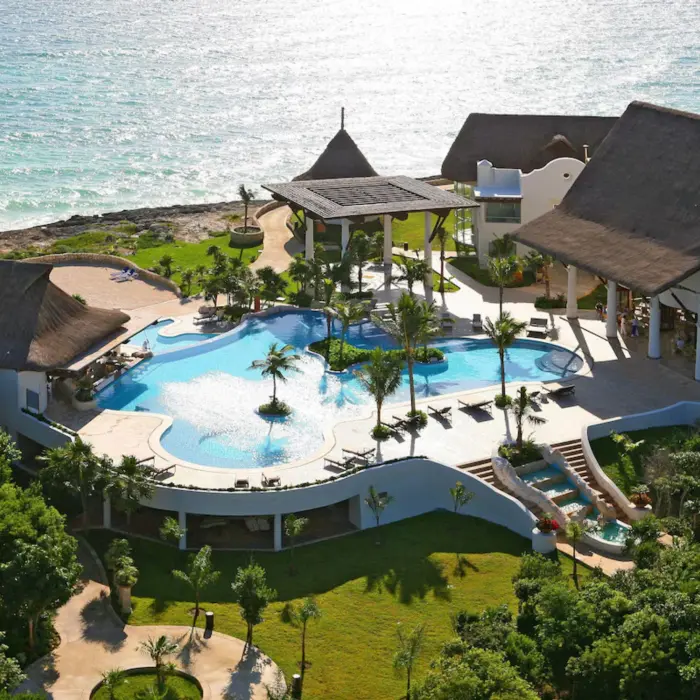 An aerial view of a resort with a large pool, lush landscaping, and buildings next to the ocean.