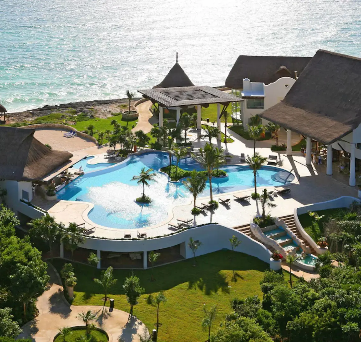 An aerial view of a resort with a large pool, lush landscaping, and buildings next to the ocean.