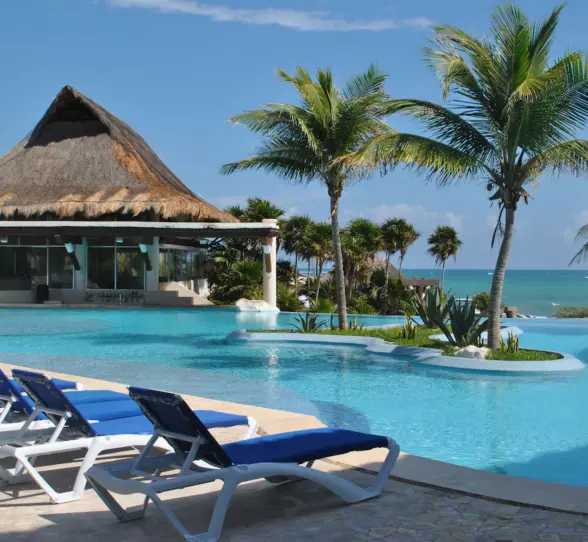 An inviting oceanfront pool area with lounge chairs, palm trees, and a traditional palapa structure.