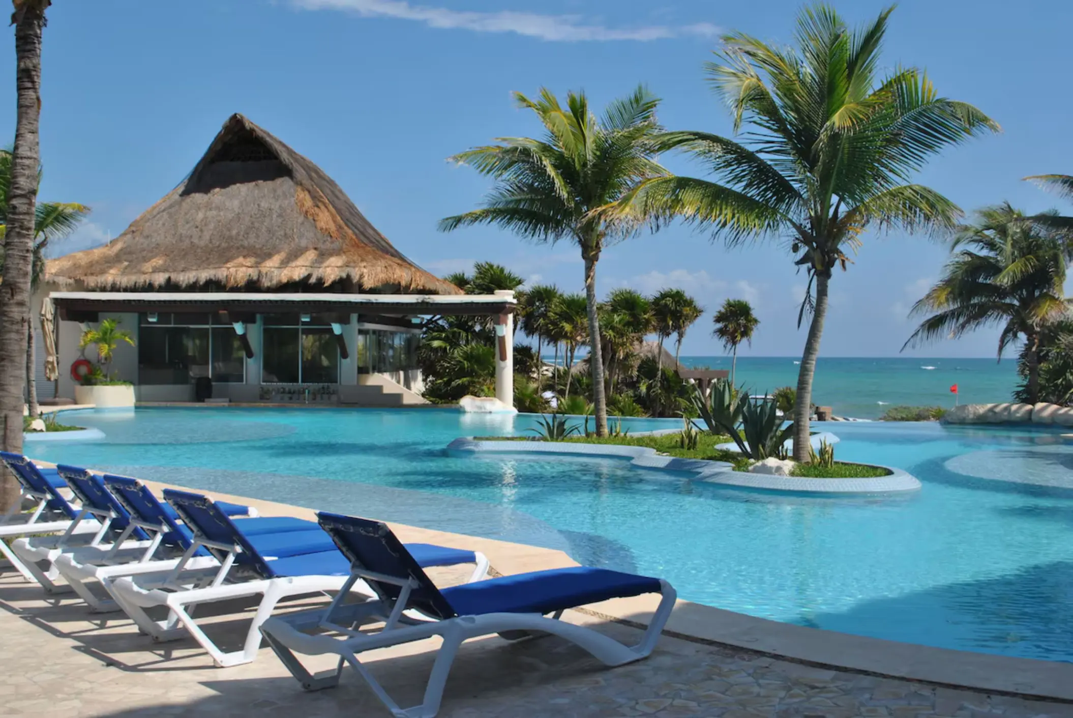 An inviting oceanfront pool area with lounge chairs, palm trees, and a traditional palapa structure.