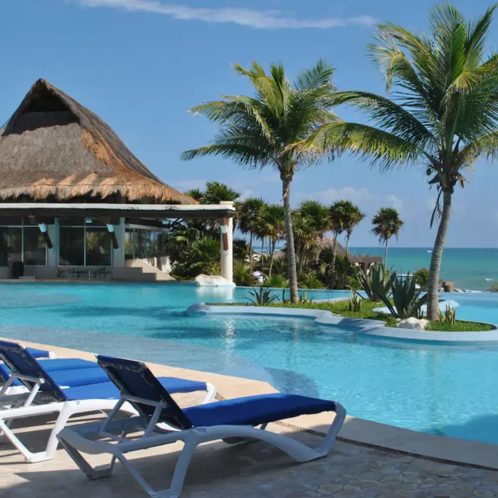 An inviting oceanfront pool area with lounge chairs, palm trees, and a traditional palapa structure.
