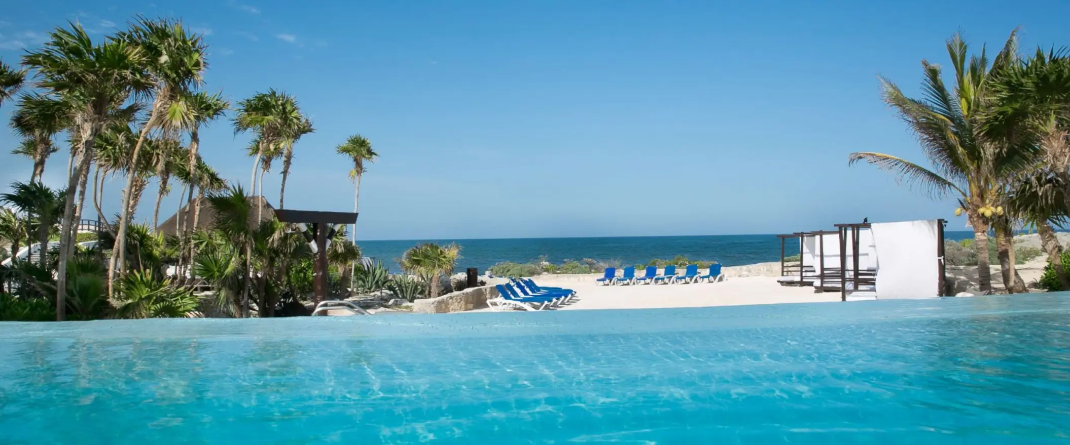An infinity pool overlooking a white sandy beach with blue lounge chairs and the clear blue ocean.