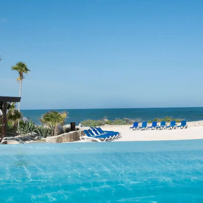 An infinity pool overlooking a white sandy beach with blue lounge chairs and the clear blue ocean.
