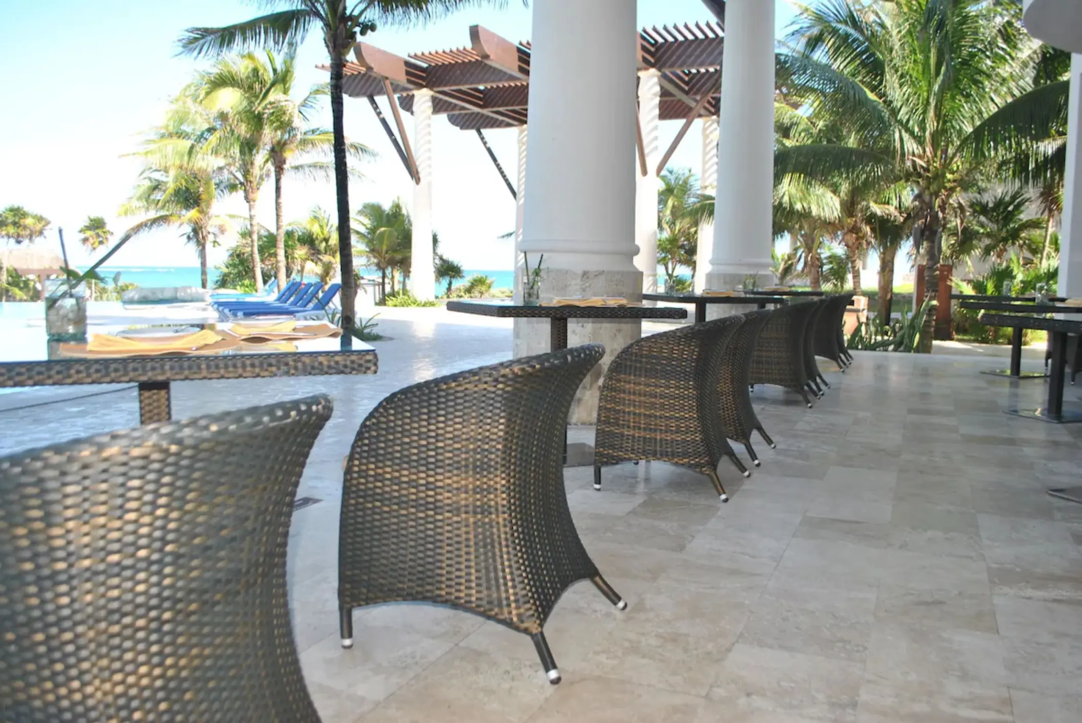 An outdoor restaurant terrace with wicker chairs and tables, surrounded by palm trees and views of the sea.