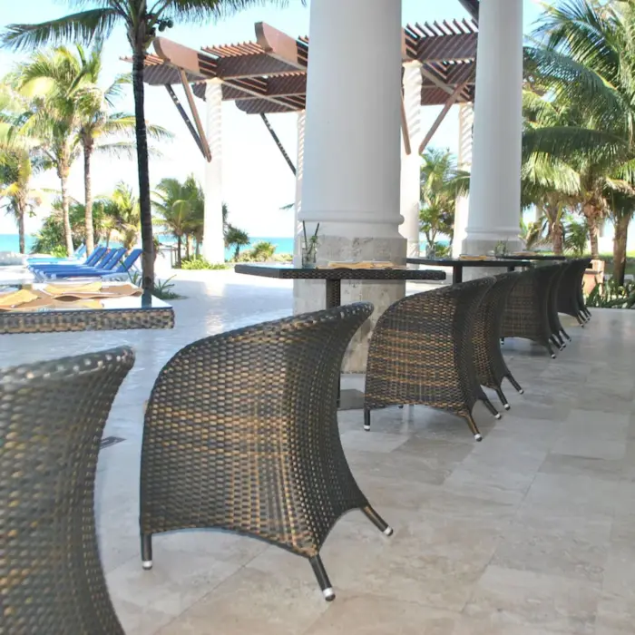 An outdoor restaurant terrace with wicker chairs and tables, surrounded by palm trees and views of the sea.