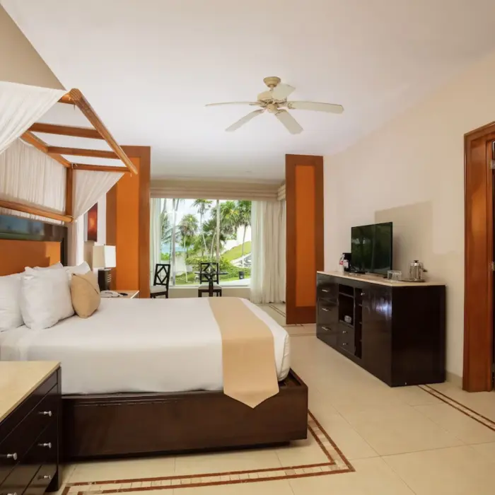 A spacious hotel suite featuring a four-poster king bed and a sitting area with a view of palm trees.