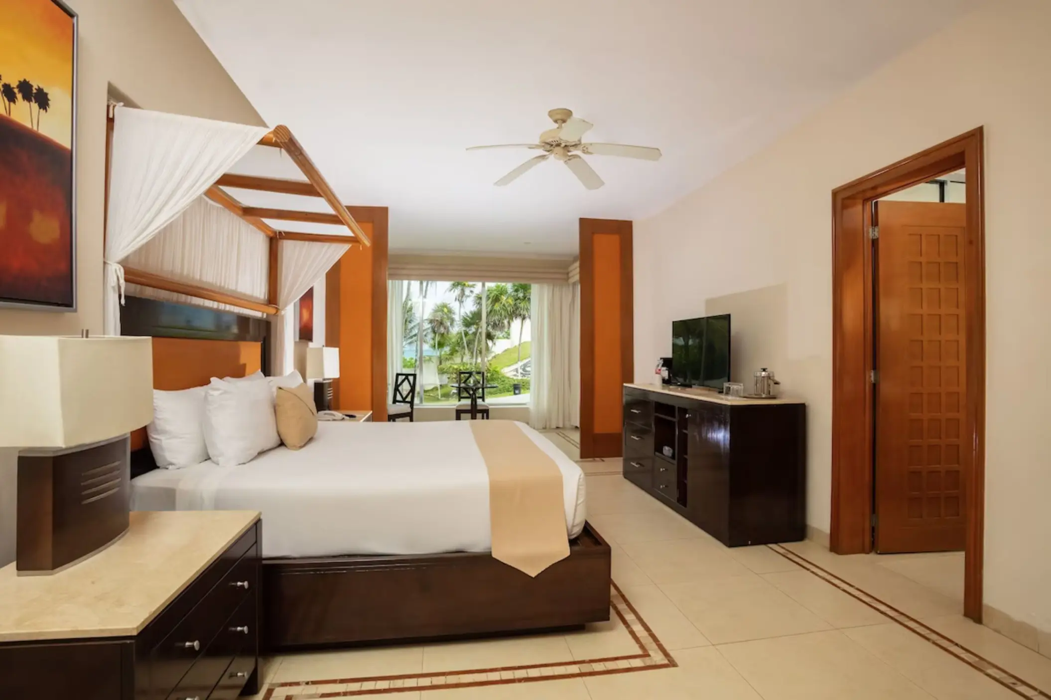 A spacious hotel suite featuring a four-poster king bed and a sitting area with a view of palm trees.