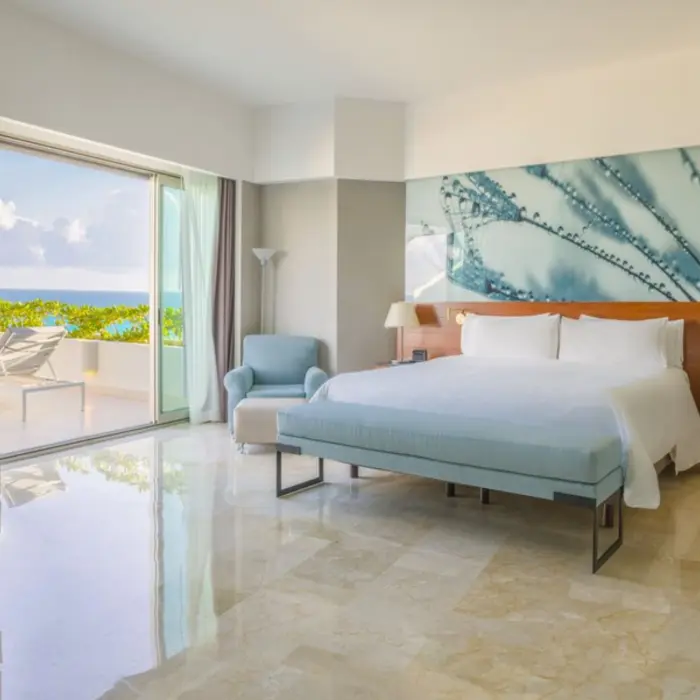 A spacious, sunlit hotel suite with a large bed, a sofa, and a balcony overlooking the ocean.