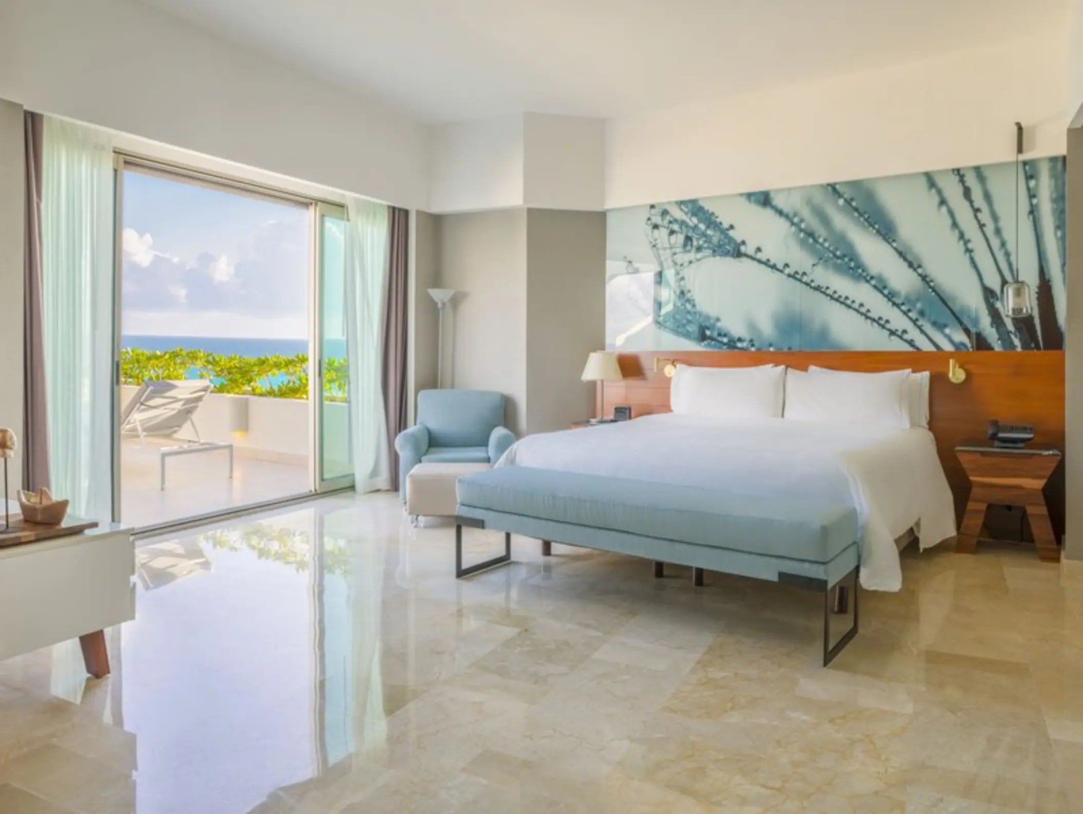 A spacious, sunlit hotel suite with a large bed, a sofa, and a balcony overlooking the ocean.
