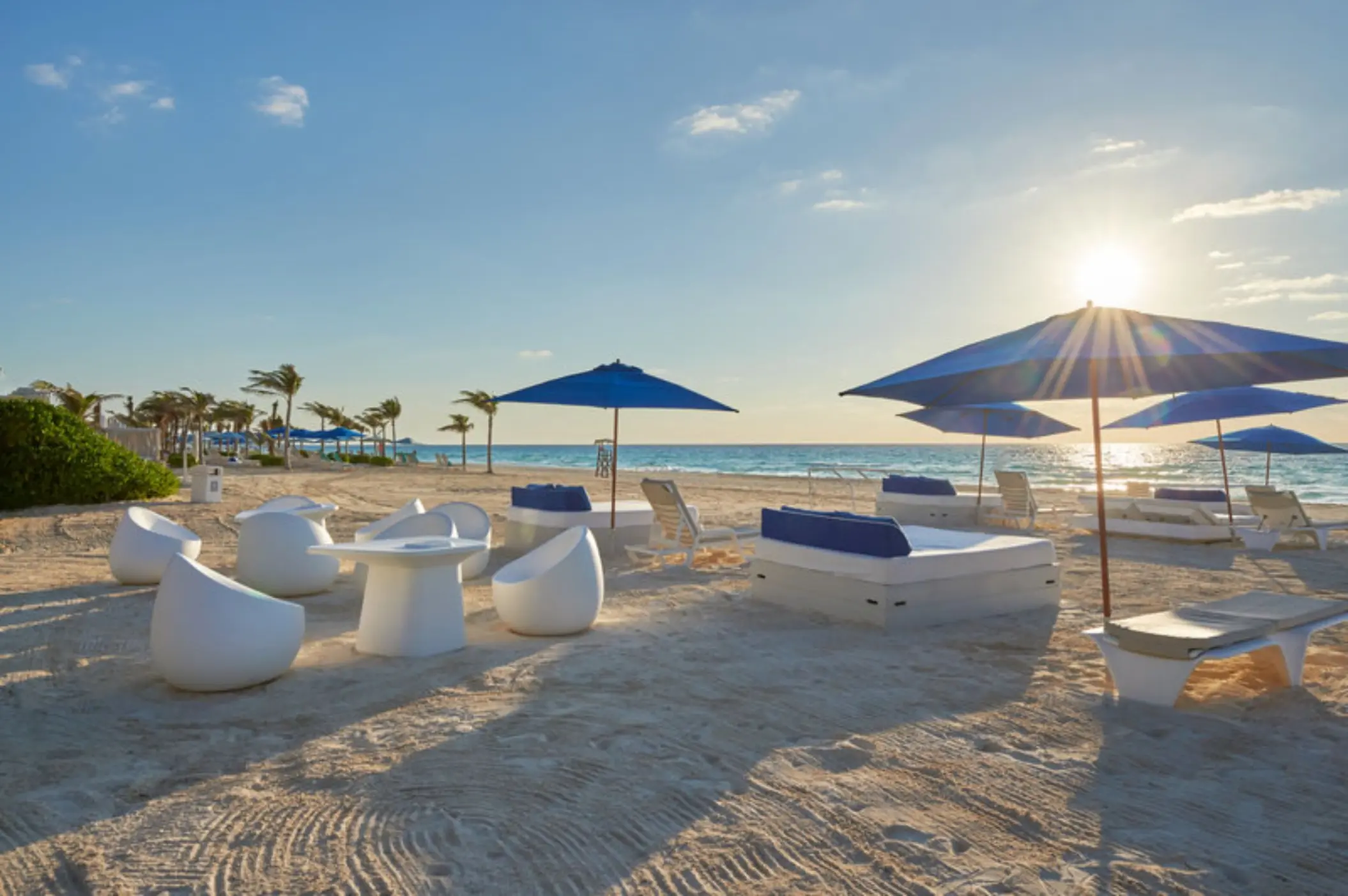 A sunny beach with white lounge chairs, blue umbrellas, and a calm, turquoise ocean in the background.