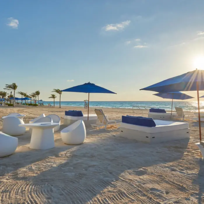 A sunny beach with white lounge chairs, blue umbrellas, and a calm, turquoise ocean in the background.