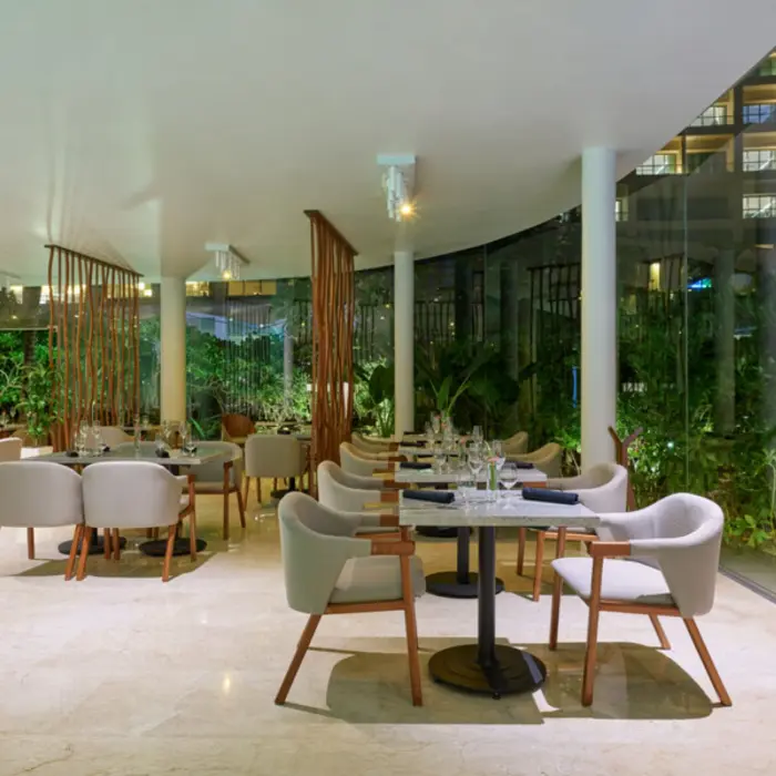A modern steakhouse at night with a large glass wall looking out onto lush, green foliage.