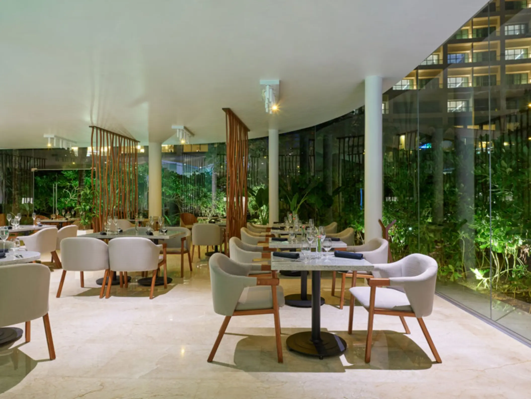 A modern steakhouse at night with a large glass wall looking out onto lush, green foliage.